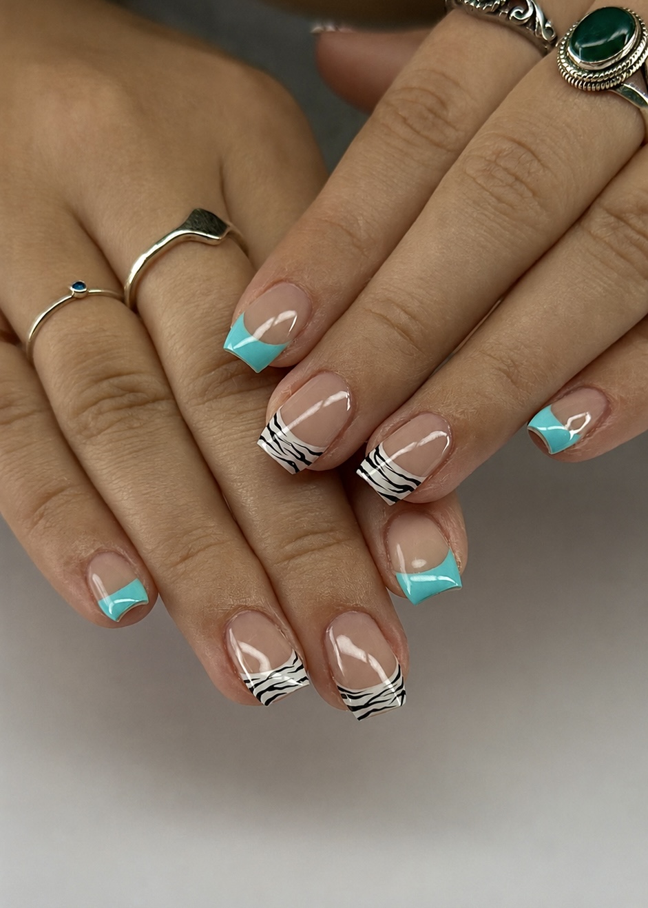 Two hands with square nails, nude bases, turquoise French tips, and black-and-white zebra accent tips